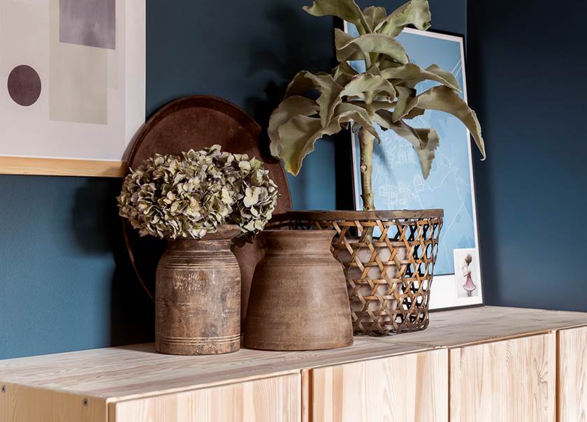 Bureau with plants and painting on it and blue colored walls in the background.