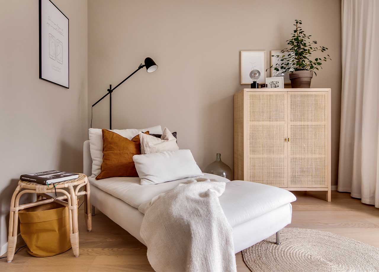 A white divan with pillows in white and brown, and a white blanket. Behind is a black floor lamp and on the side there is a small table in rattan.